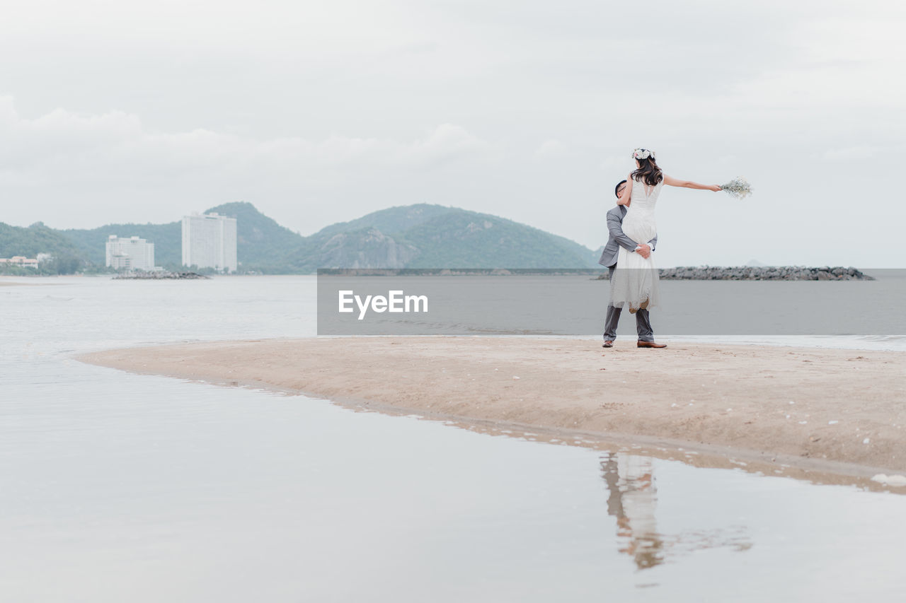 Newlywed couple standing on beach against sky