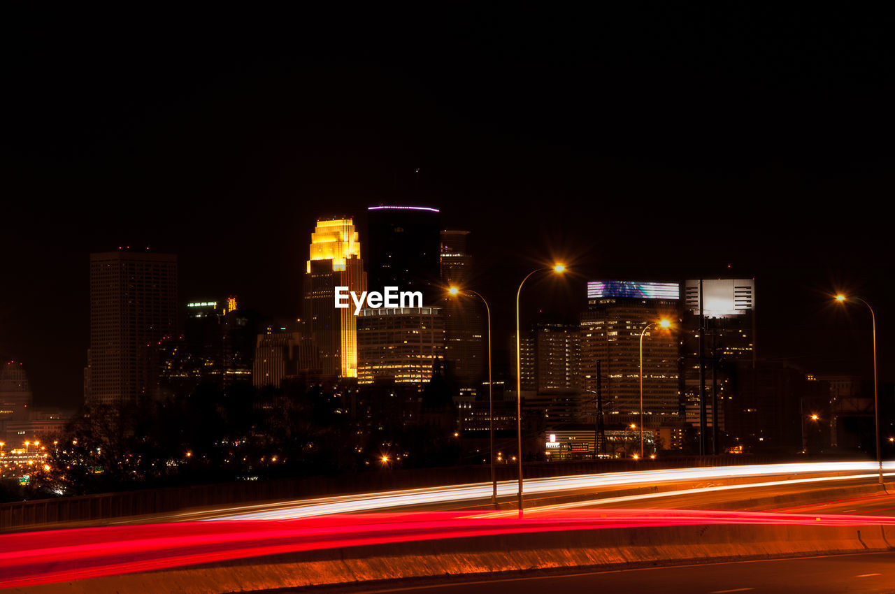 ILLUMINATED LIGHT TRAILS ON ROAD AGAINST BUILDINGS AT NIGHT