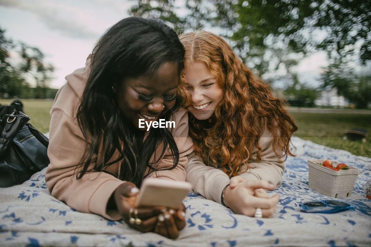 Happy teenage girl lying beside female friend using smart phone on picnic blanket