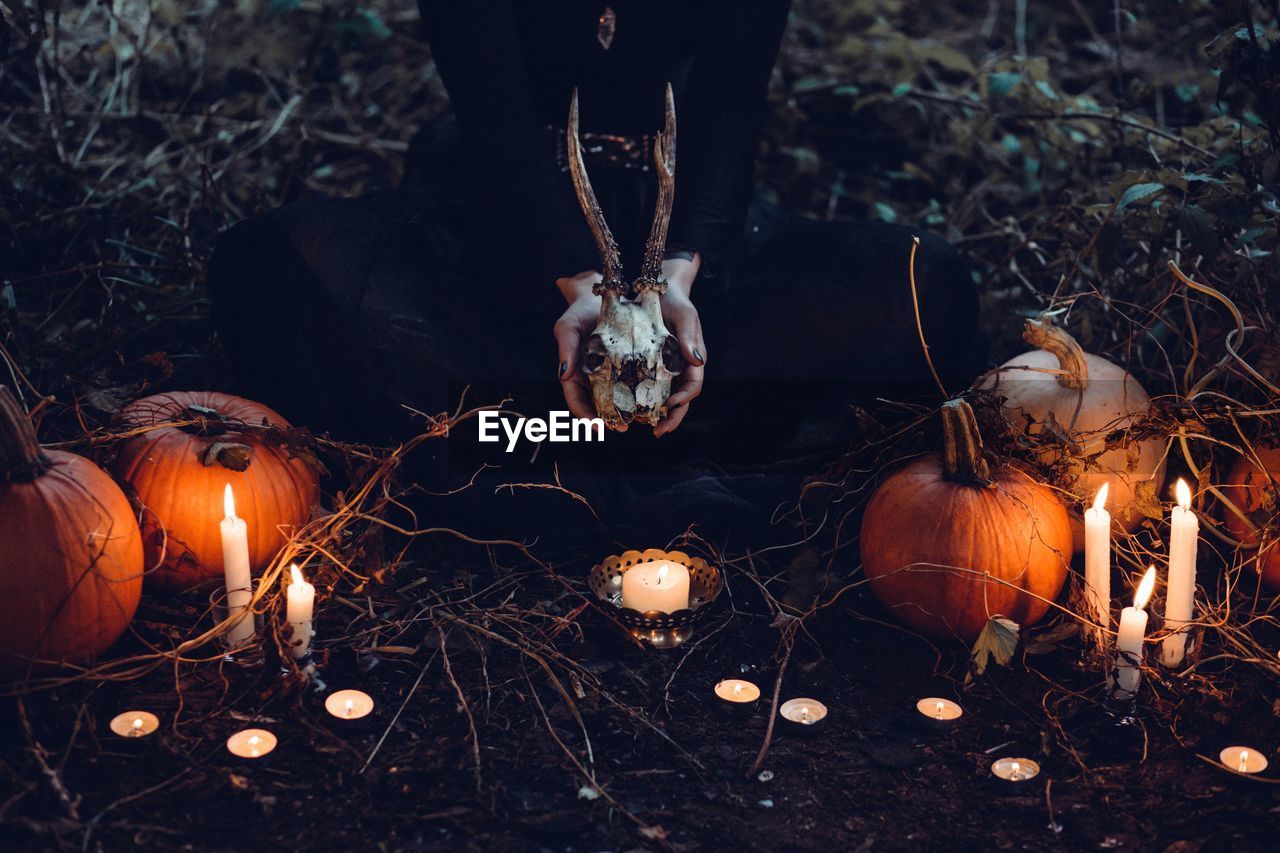Low section of woman holding animal skull while sitting amidst pumpkins and burning candles during halloween