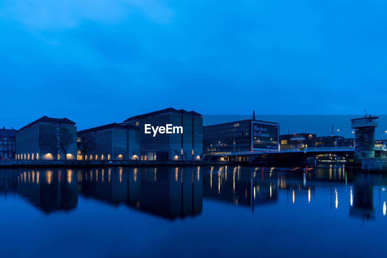 Illuminated buildings by river against sky at dusk