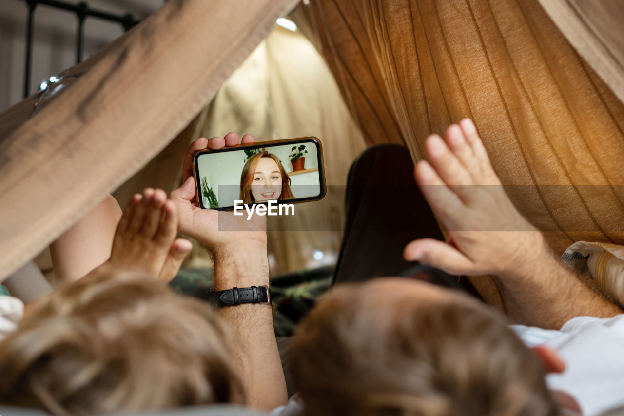 Father and son using mobile phone while lying in children's tent, making video call with family.