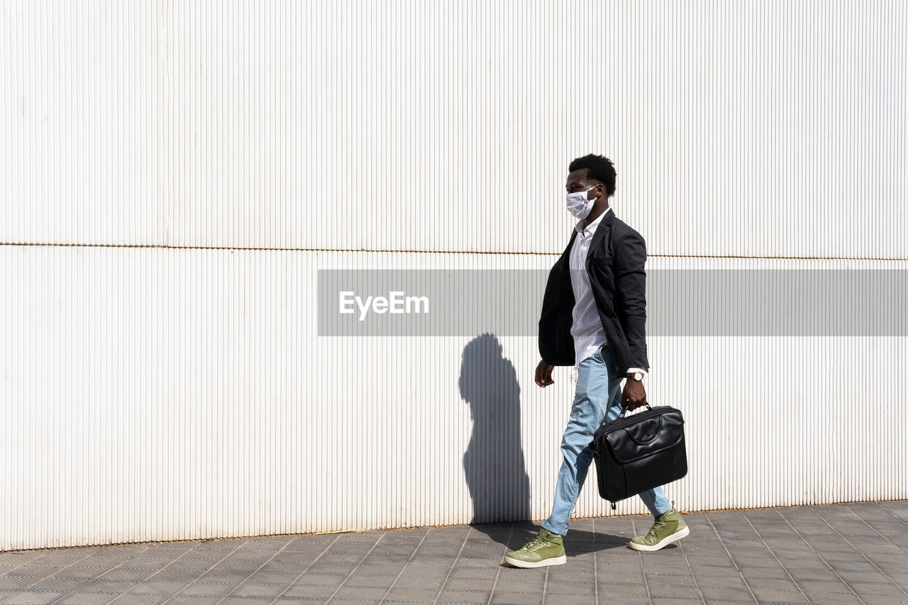 Side view of well dressed black male entrepreneur in medical mask walking along urban building on sunny day in city during coronavirus epidemic