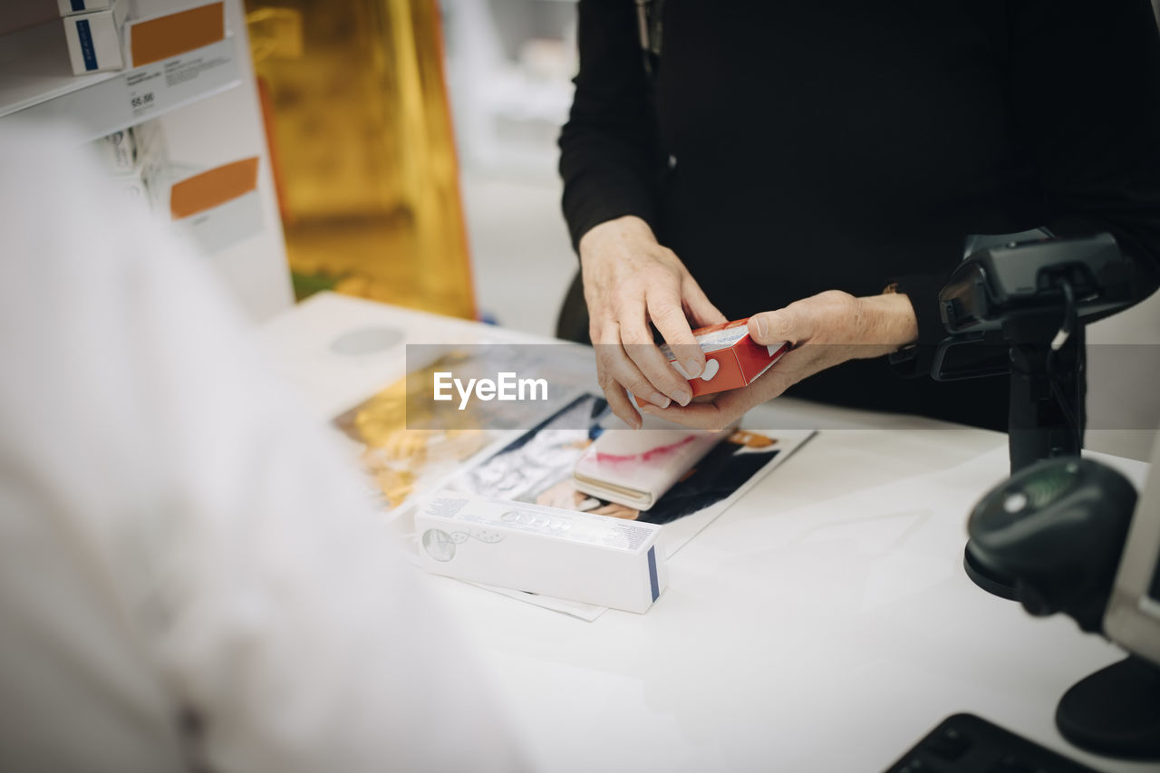 Midsection of senior customer holding prescription medicine with female owner at checkout in pharmacy store