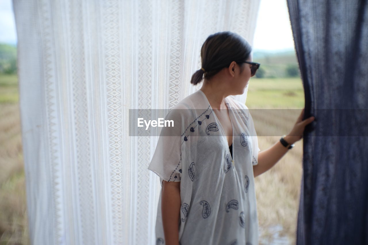 Woman holding curtain while looking through window at home