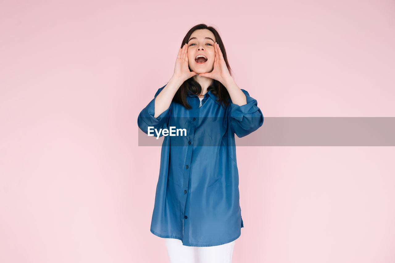 one person, studio shot, indoors, women, clothing, emotion, adult, pink, colored background, portrait, copy space, front view, blue, young adult, outerwear, female, happiness, facial expression, standing, hand, sleeve, smiling, dress, eyes closed, long hair, mouth open, hairstyle, three quarter length
