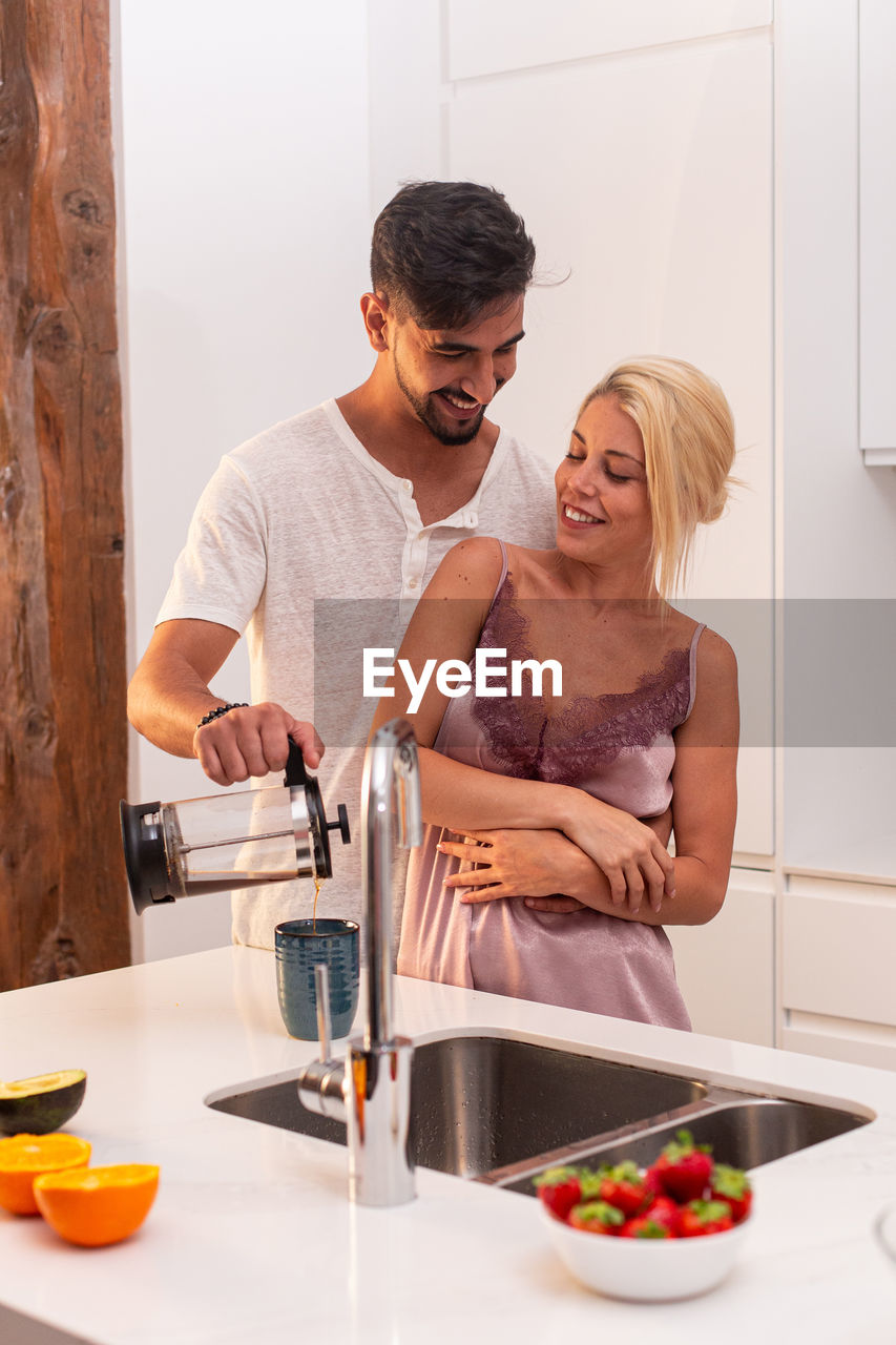 Loving couple in pajamas hugging and pouring fresh coffee in cup together while standing at table in kitchen during breakfast in morning