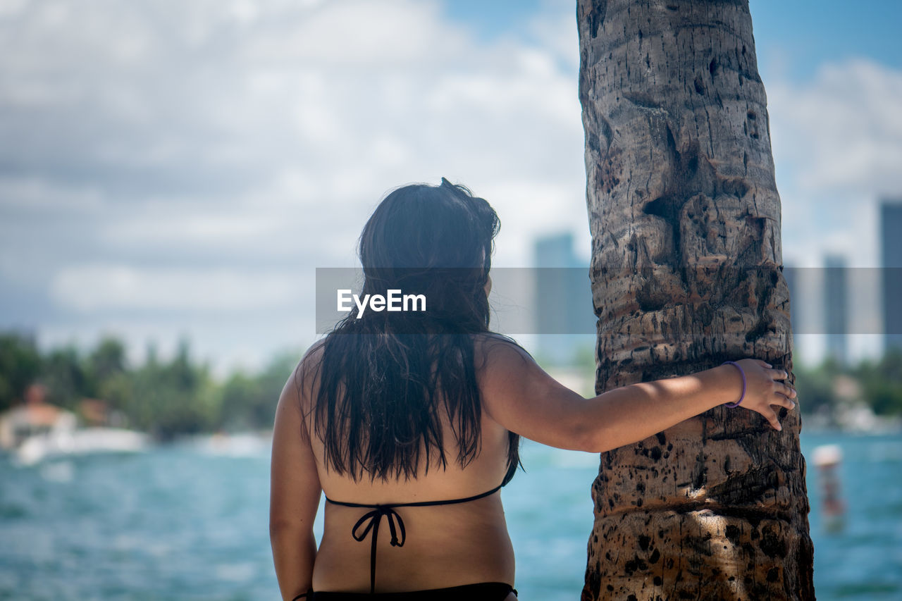 Rear view of woman standing by tree trunk against sky