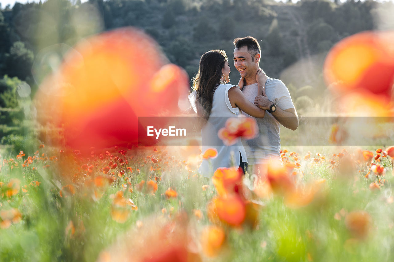 Happy romantic couple smiling at each other while standing on poppy flower field
