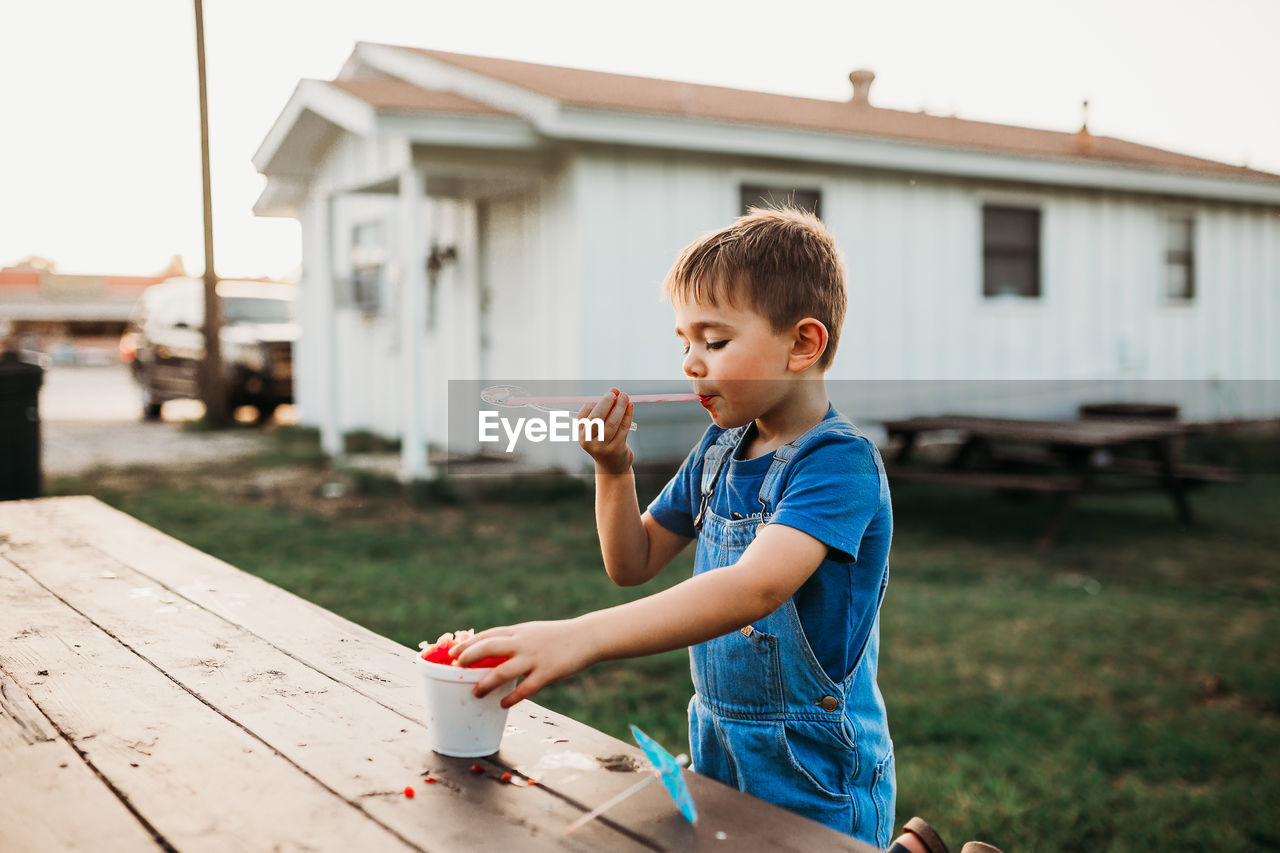 Young boy sitting at picnic table eating a red snow cone at sunset