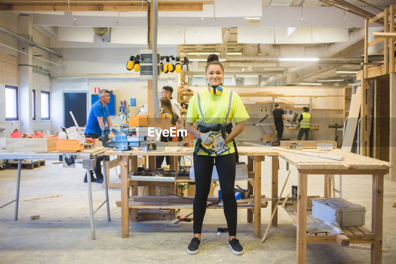 Full length portrait of smiling young female trainee holding power tool while standing by workbench at illuminated works