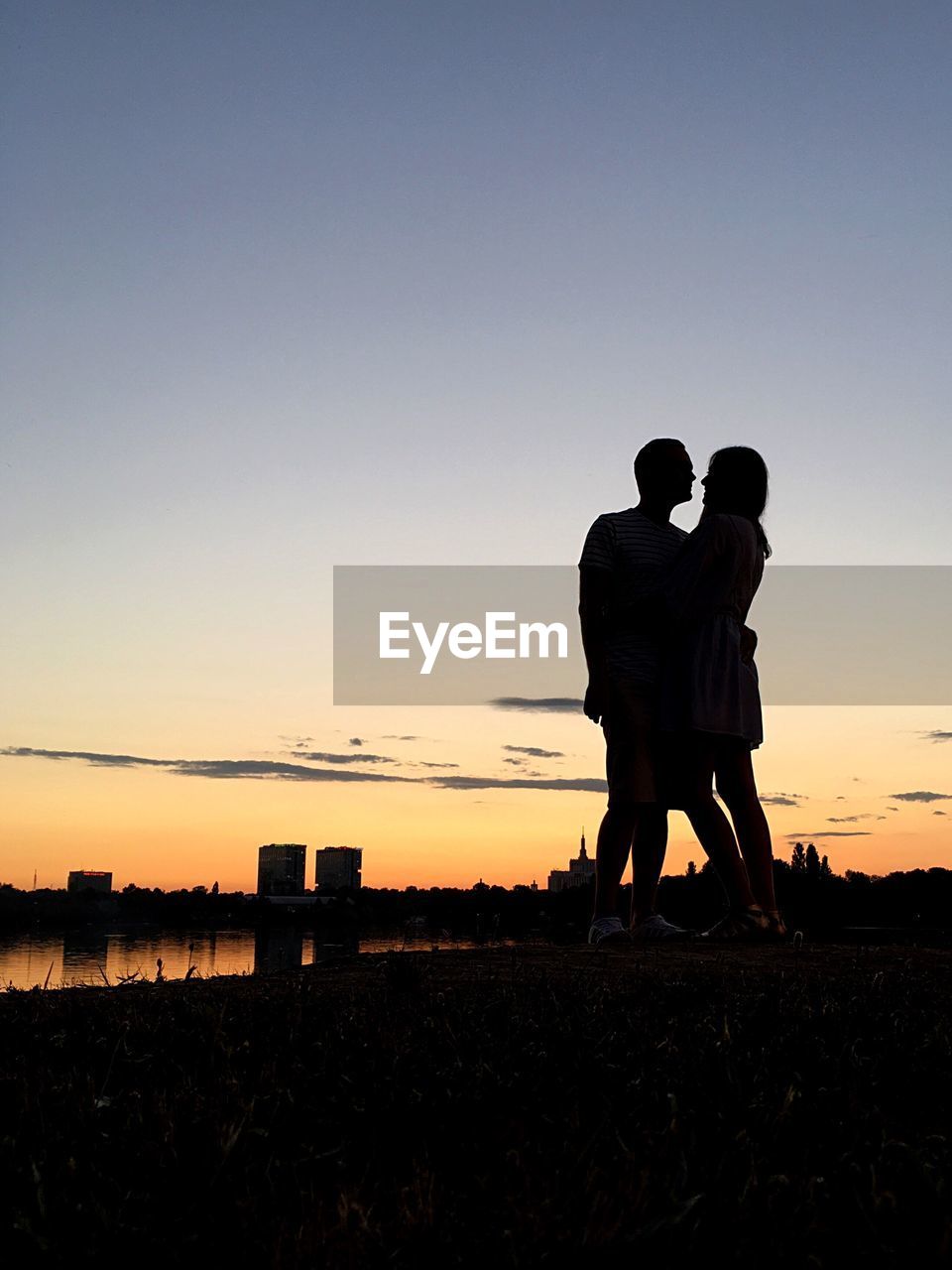 Couple standing at lakeshore against sky during sunset