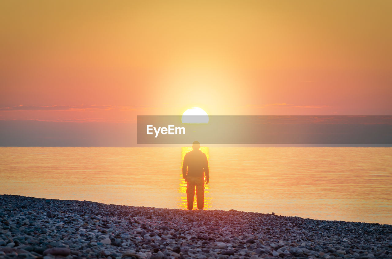 FULL LENGTH OF WOMAN STANDING ON SEA SHORE AGAINST ORANGE SKY