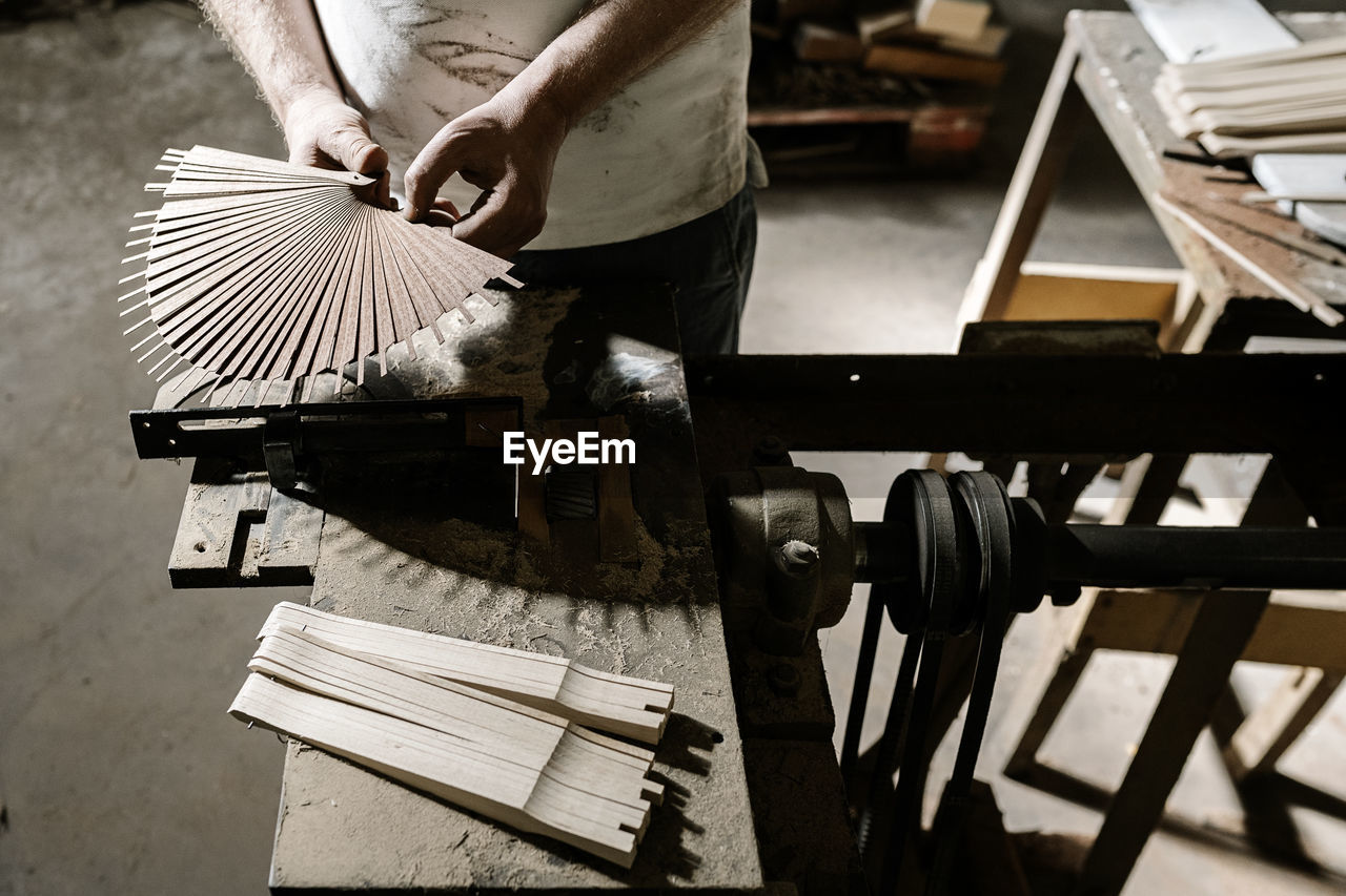 From above of unrecognizable crop craftsman working in dirty workshop at dusty workbench while standing with wooden folding fan