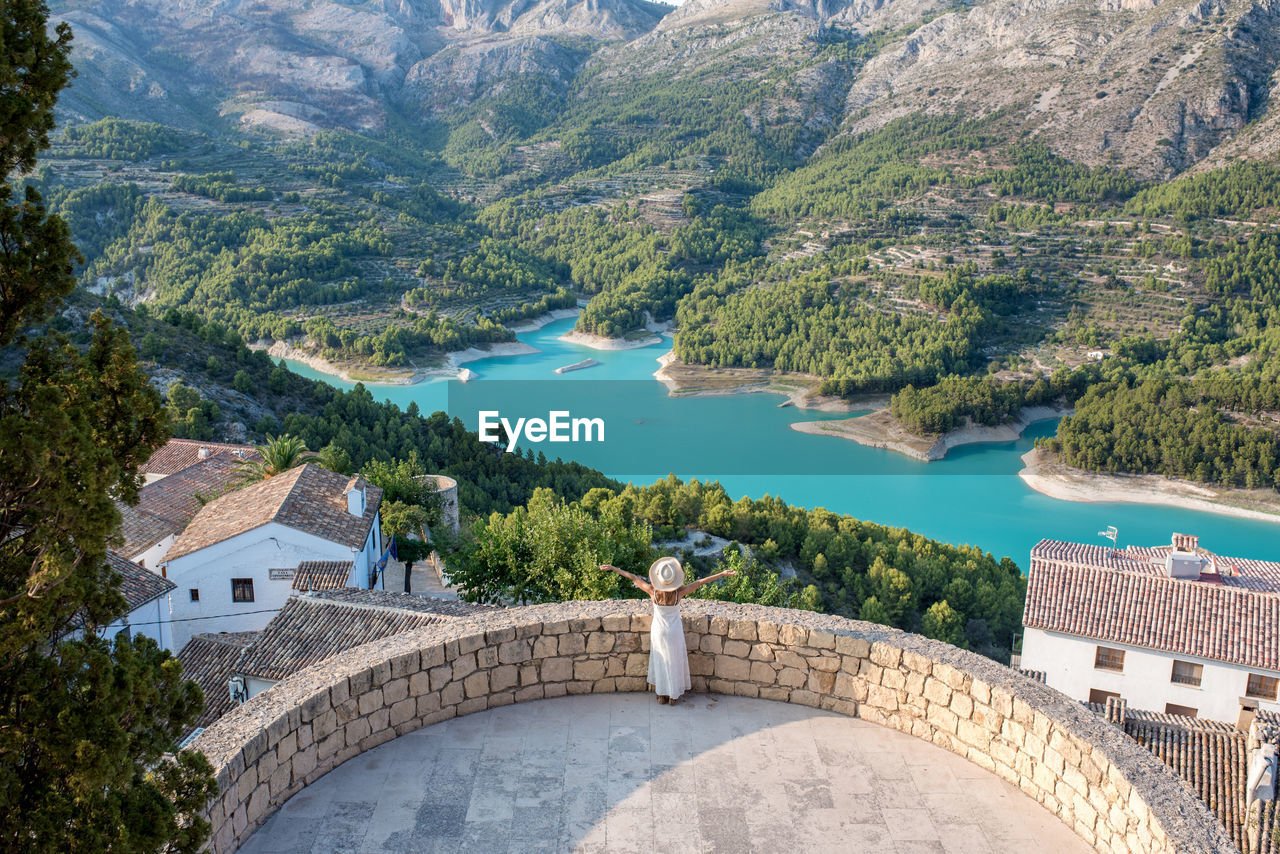 From above back view of distant unrecognizable female traveler standing near stone fence above town with aged rooftops while admiring lake surrounded by mountains in spain