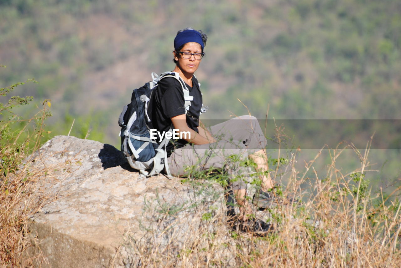 one person, nature, adult, plant, leisure activity, activity, smiling, grass, adventure, happiness, lifestyles, wilderness, clothing, outdoors, land, young adult, sitting, hiking, portrait, day, emotion, men, full length, relaxation, casual clothing, landscape, copy space, recreation, travel, looking at camera, environment, sports