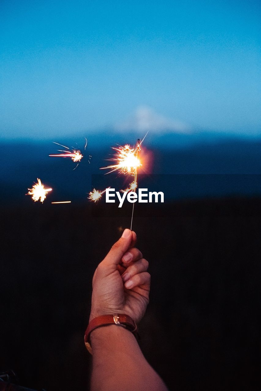 Cropped image of hand holding sparkler on field against sky at dusk