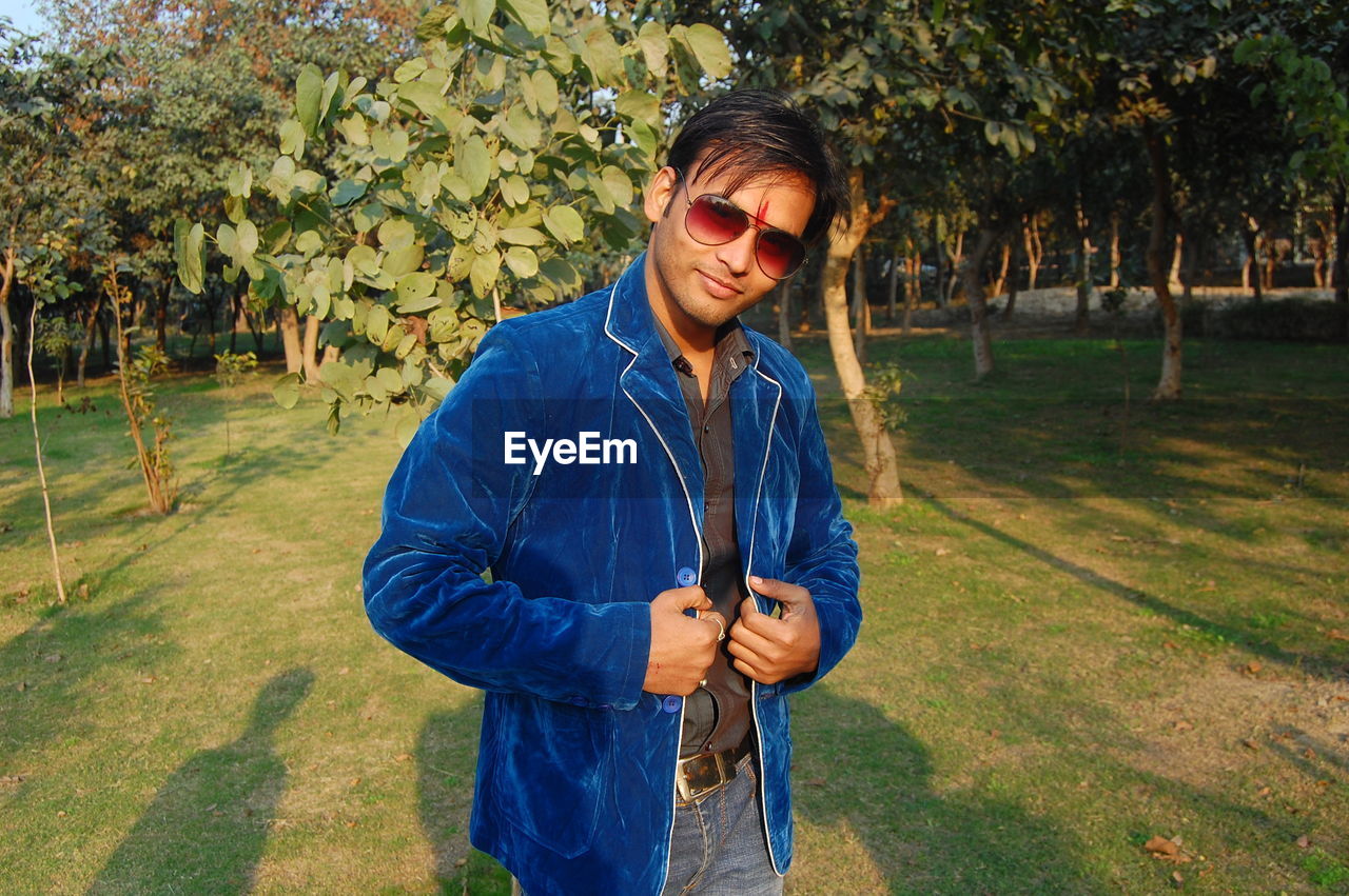Smiling man wearing sunglasses and blue jacket standing on grassy field against trees at park
