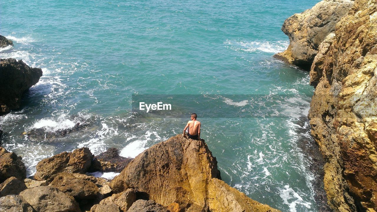 High angle view of shirtless man sitting on rock by sea