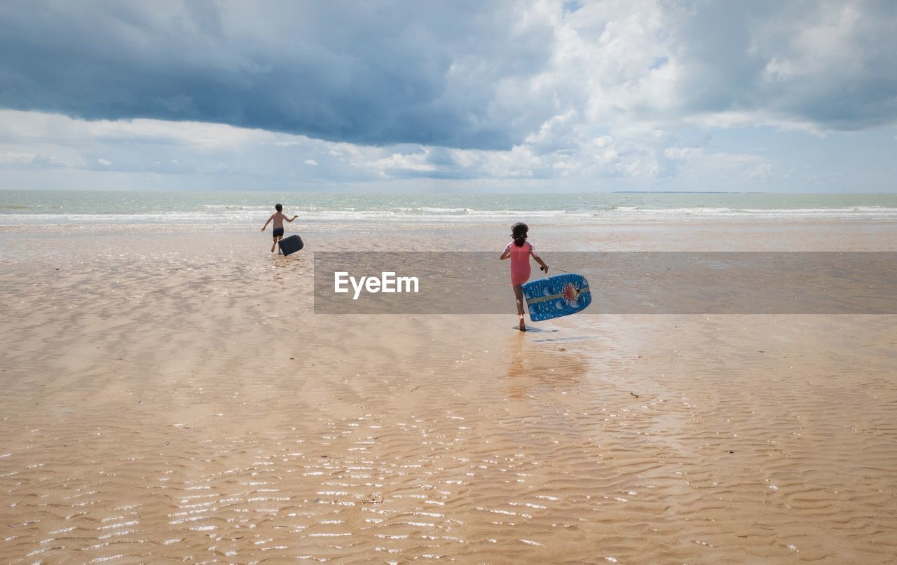 Rear view of children holding pool rafts while running towards sea against cloudy sky