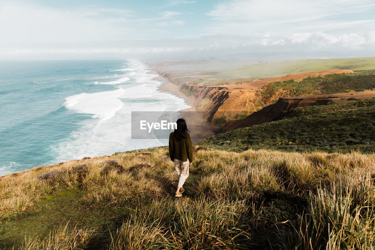 Back view of woman standing in solitude on high cliff meadow of point reyes national seashore observing majestic view of ocean in california