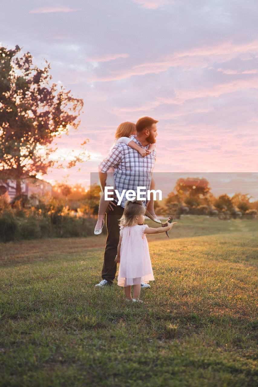 Father and daughters on grass against sky during sunset