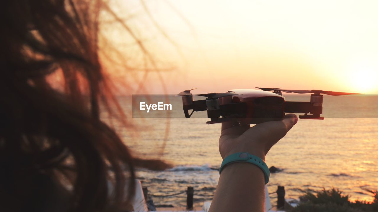 Cropped hand of woman holding drone against sea during sunset