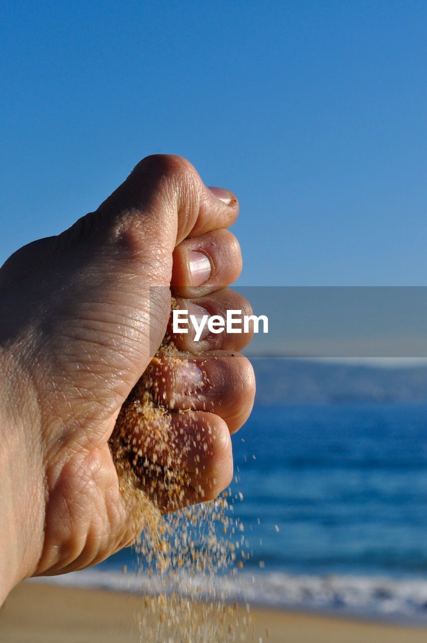 Close-up of hand holding sand against sea and clear blue sky