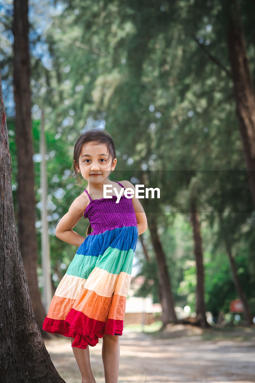 Pretty little girl wearing colourful dress and standing near the beach trees.