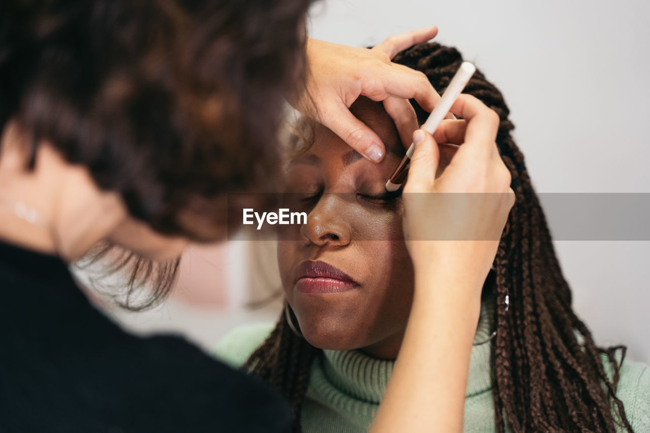 Cropped unrecognizable female visagiste spreading eyeshadow on eyelid of happy black woman during makeup session in salon