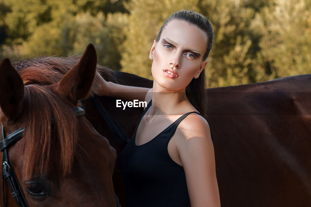 Portrait of young woman standing by brown horse on field