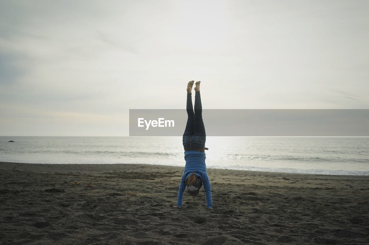 Woman performing headstand on beach against cloudy sky