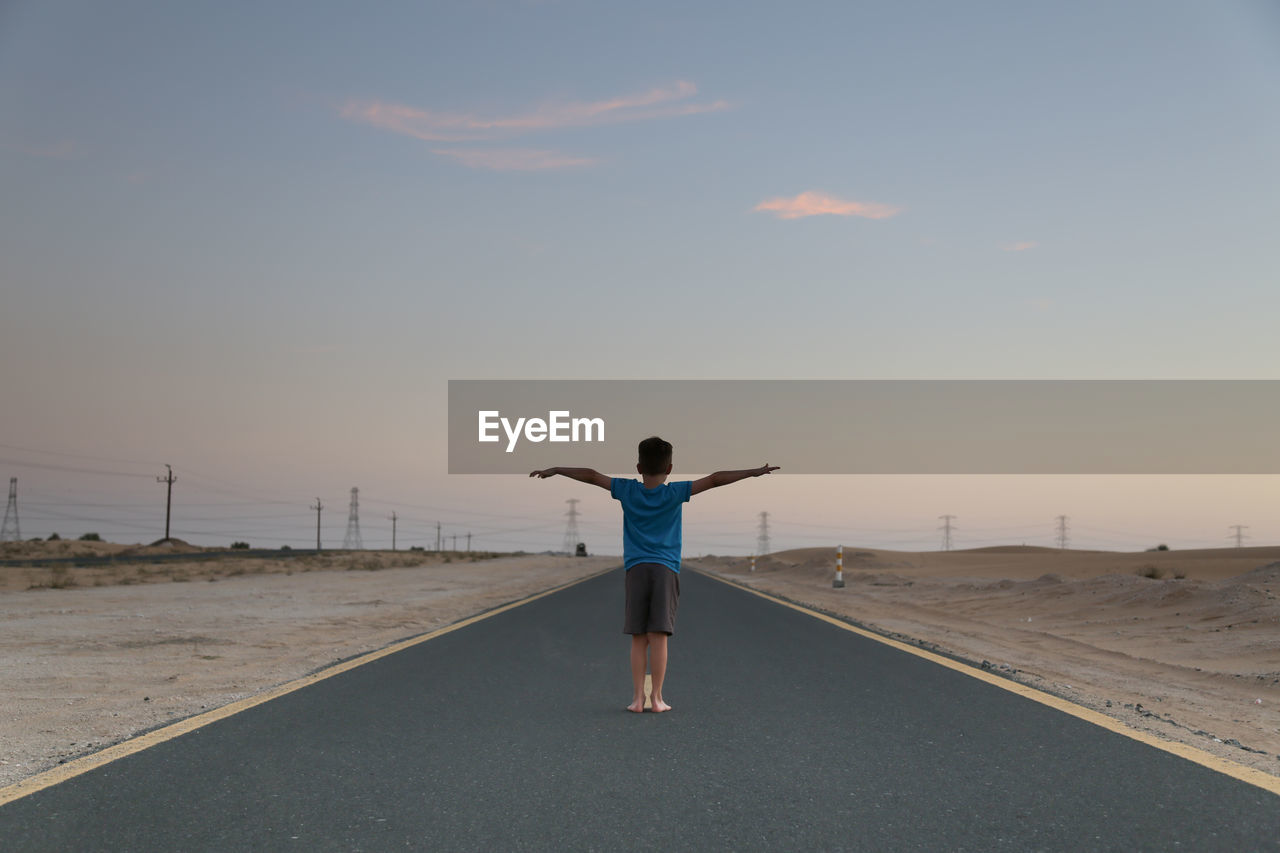 Rear view of boy with arms outstretched standing on road against sky during sunset