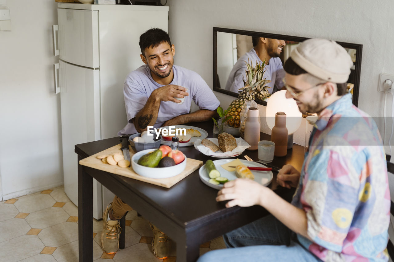 Happy gay couple having fruits while sitting at dining table in kitchen