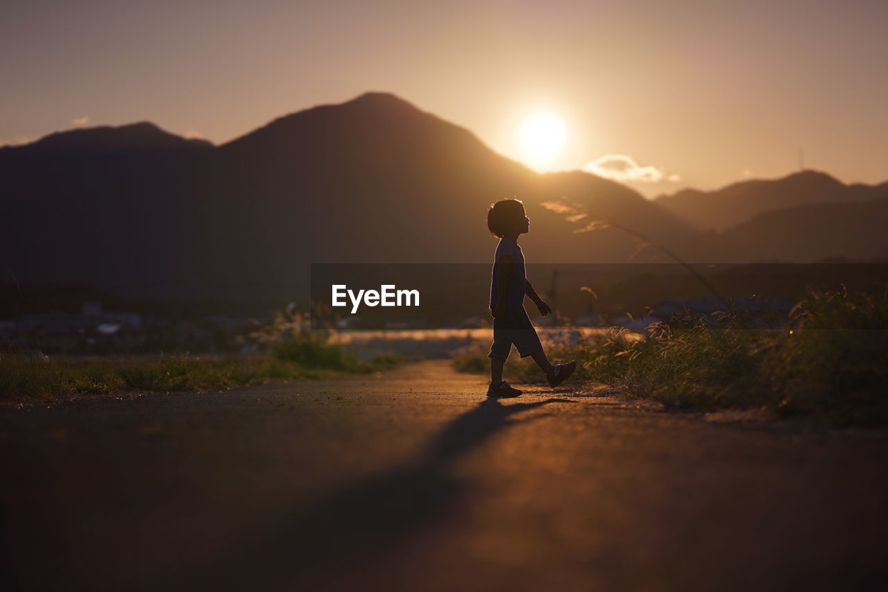 Silhouette boy walking on road towards mountain against sky during sunset