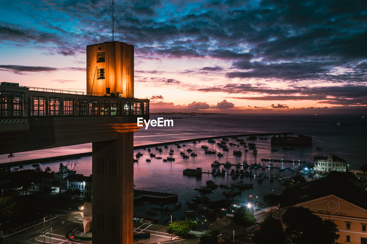 Illuminated building by sea against sky at sunset