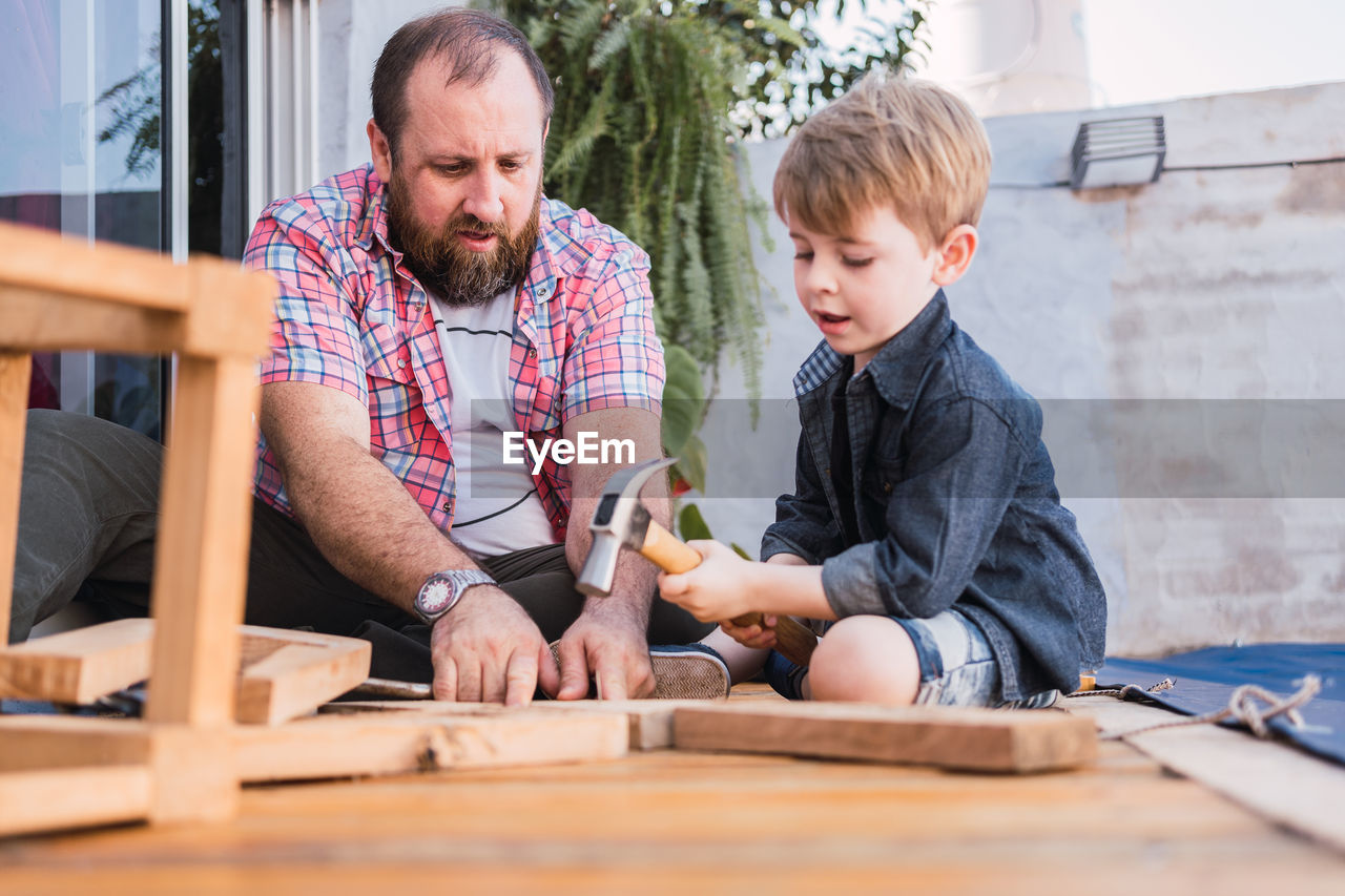 Bearded dad teaching son with hammer working with wood while sitting on boardwalk on weekend