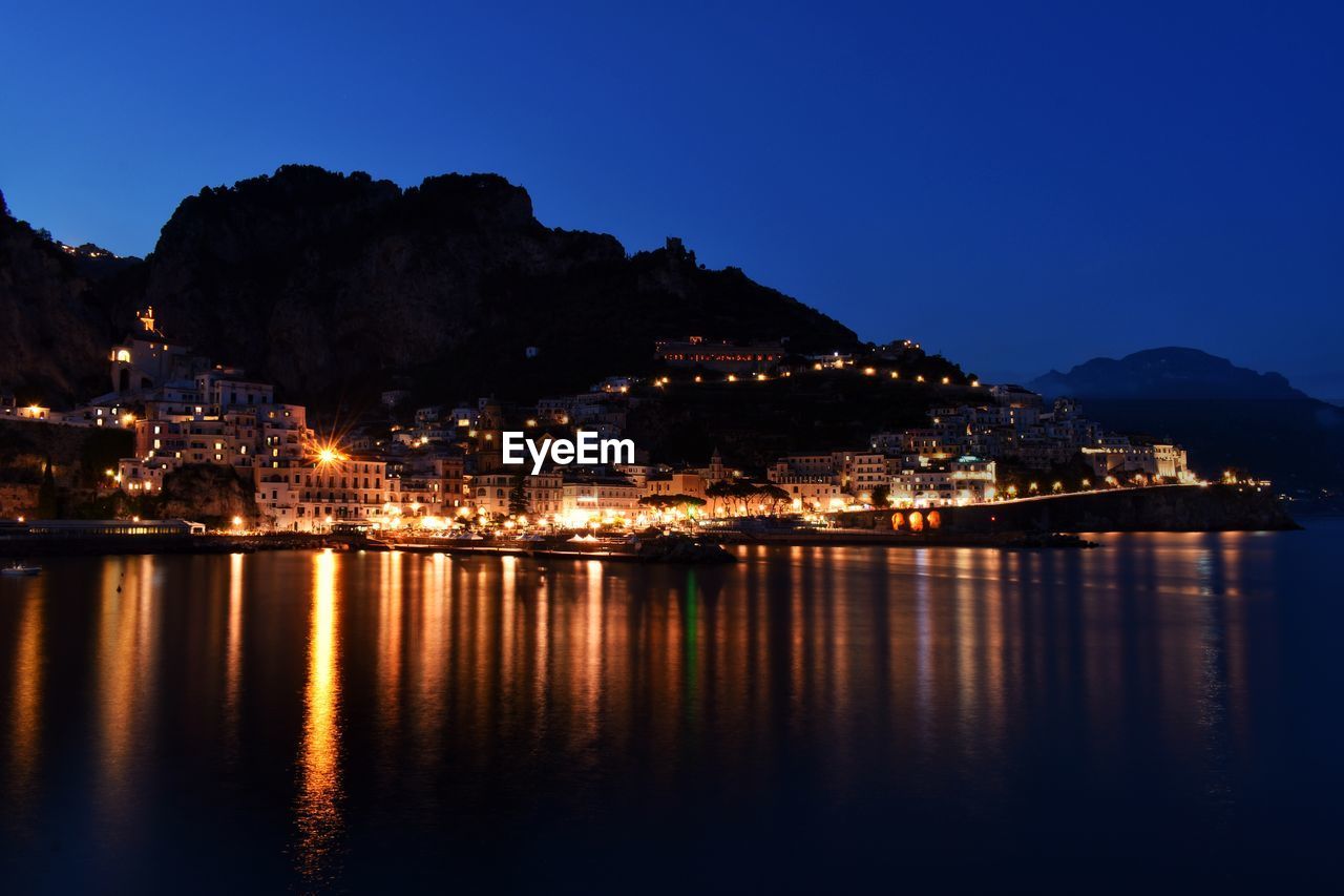 ILLUMINATED CITY BY SEA AGAINST SKY AT NIGHT