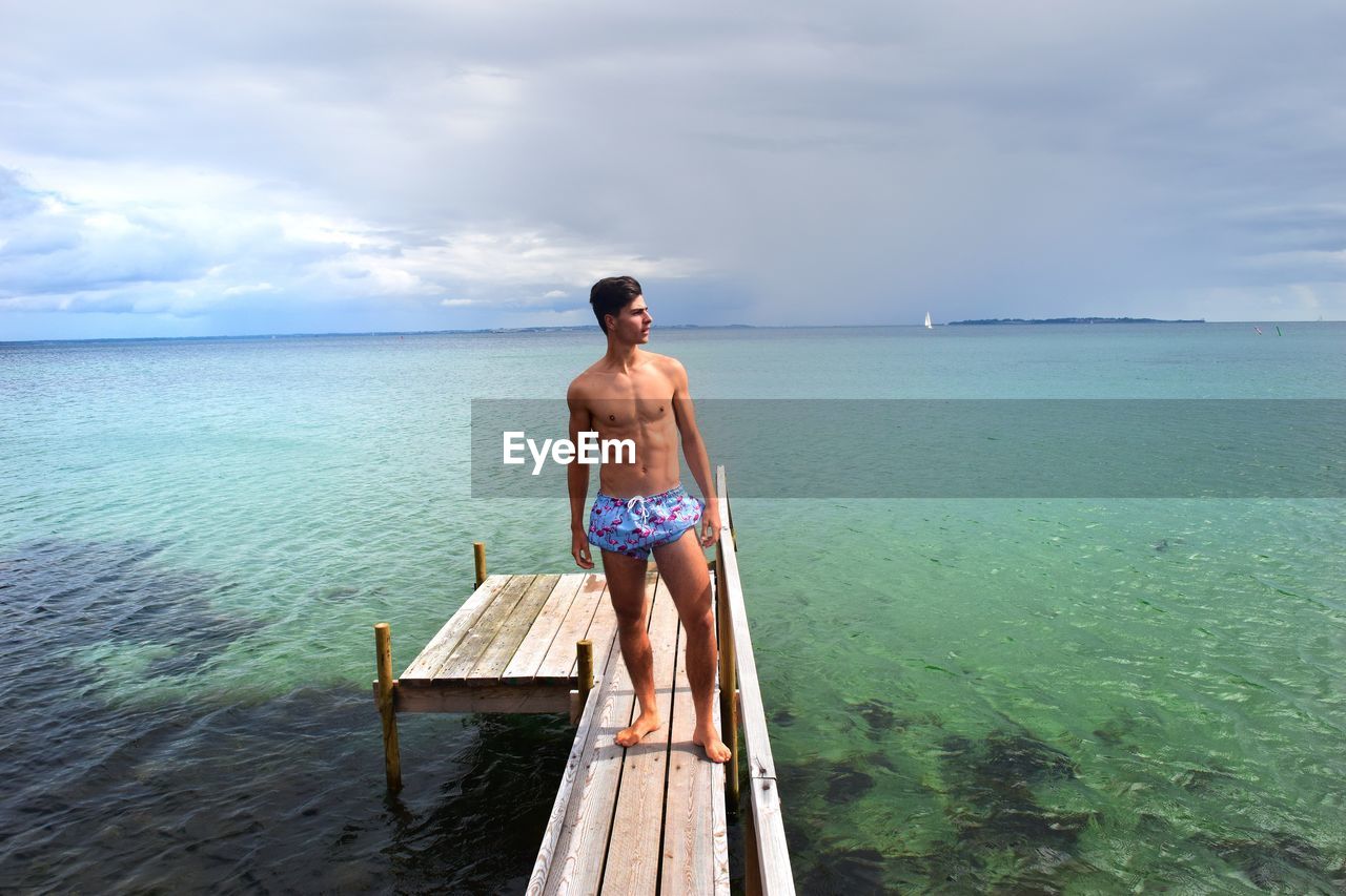 Full length of shirtless man standing on pier over sea against sky