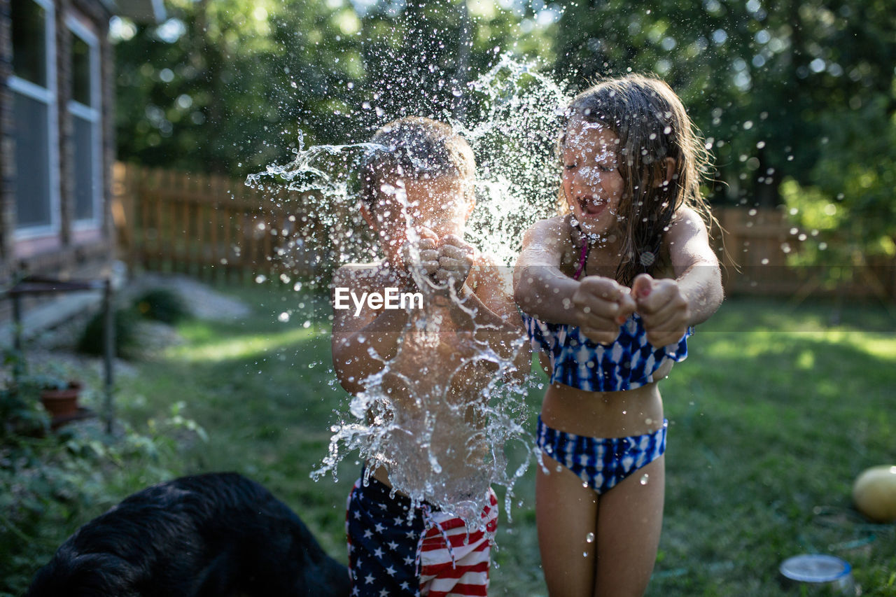 Cheerful siblings bursting water bombs while standing at backyard