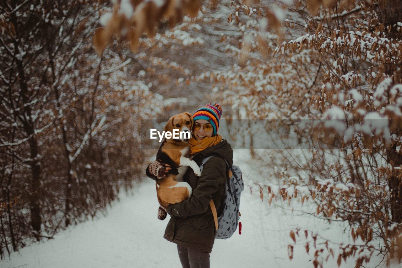 Portrait of smiling young woman carrying dog while standing in forest during winter