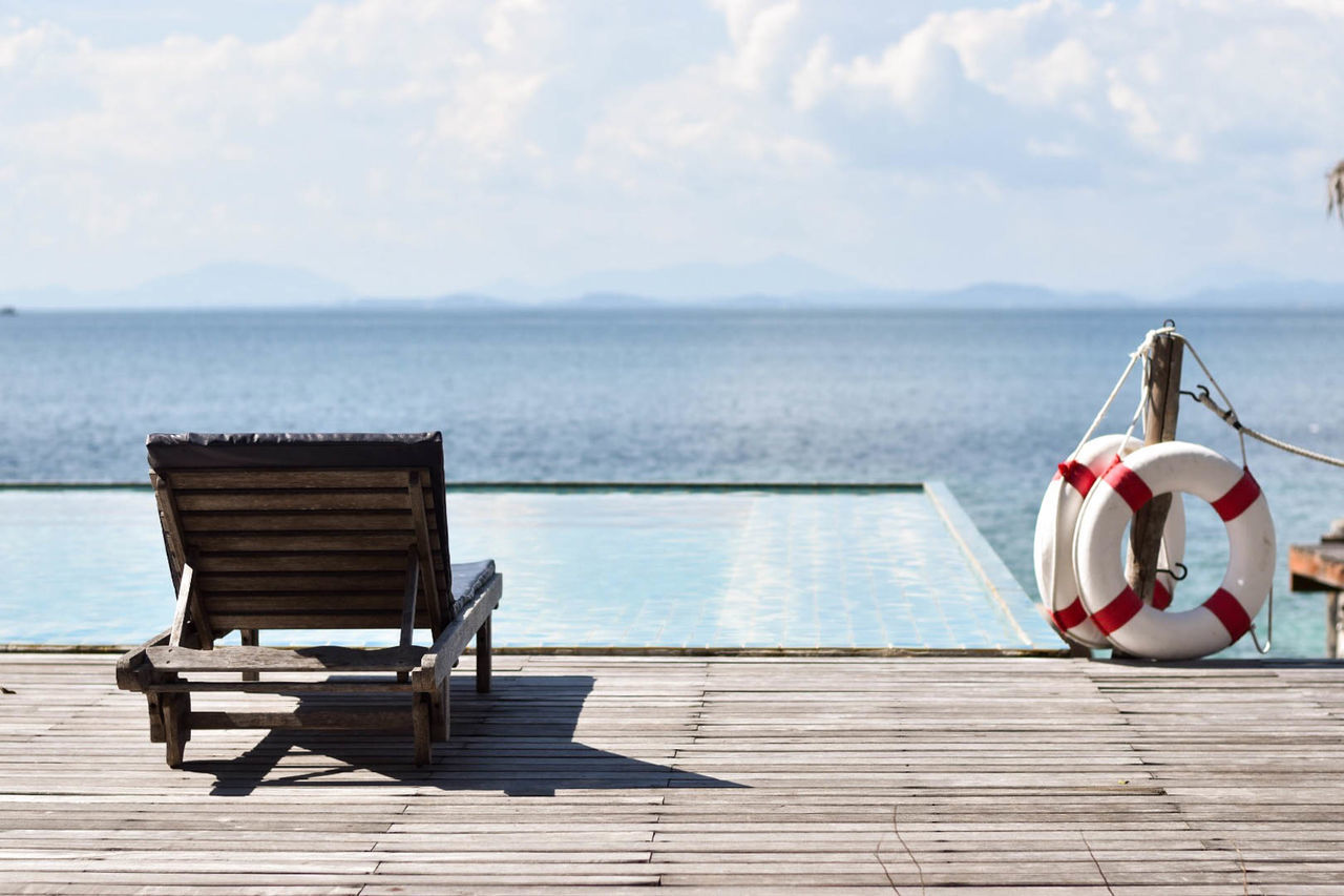 Empty lounge chair by swimming pool at tourist resort against sea on sunny day