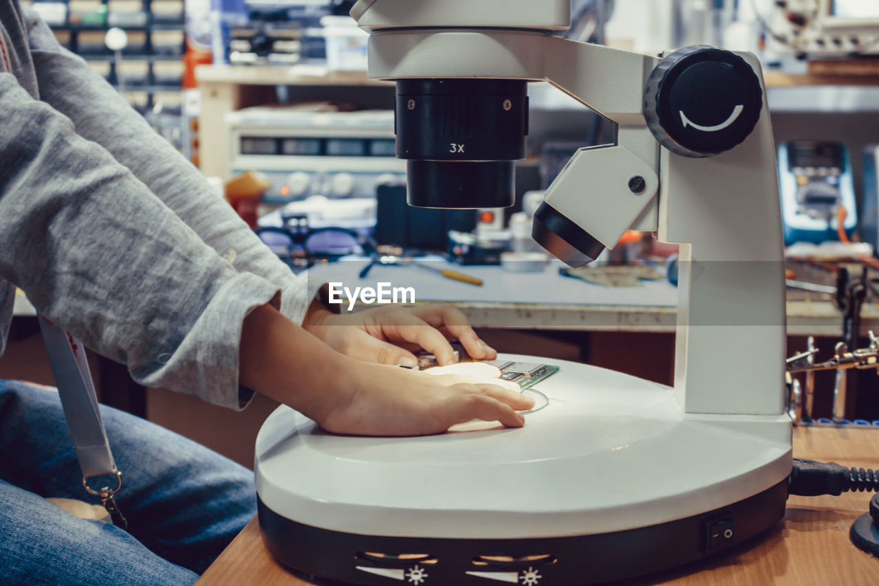 Unrecognizable kid using microscope while examining circuit board in engineering lab.