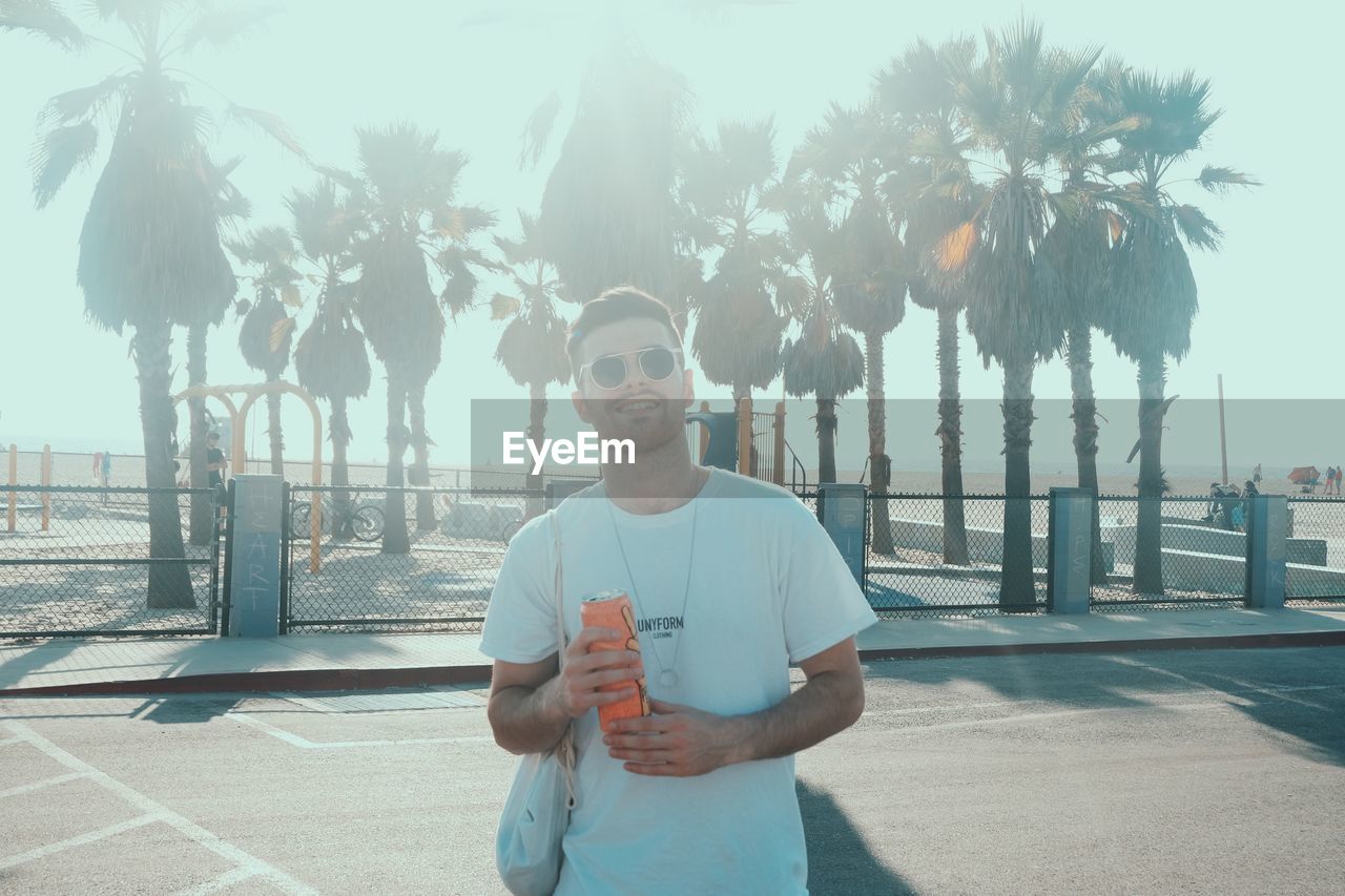 PORTRAIT OF SMILING MAN STANDING BY PALM TREE AGAINST SKY