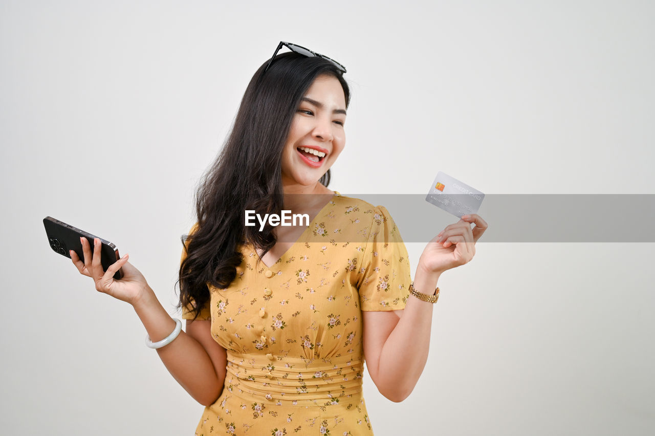 smiling, happiness, women, adult, one person, wireless technology, communication, technology, studio shot, smartphone, holding, mobile phone, indoors, portable information device, young adult, cheerful, emotion, photo shoot, portrait, long hair, smile, waist up, teeth, person, clothing, fashion, hairstyle, telephone, white background, front view, fun, positive emotion, lifestyles, finance, standing, enjoyment, looking, sleeve, casual clothing, credit card, laughing, activity, hand, copy space