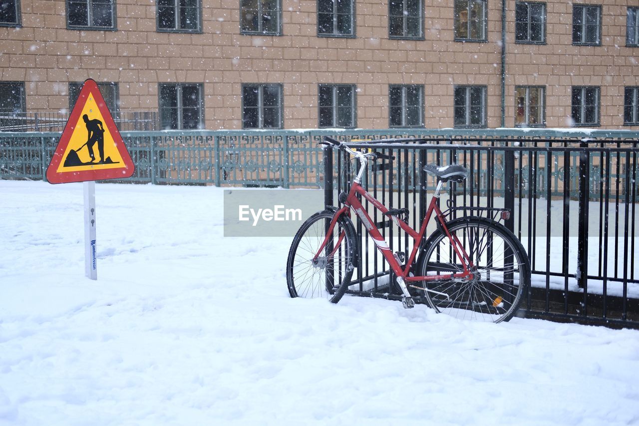 snow, cold temperature, bicycle, winter, architecture, transportation, built structure, building exterior, sign, vehicle, nature, city, communication, no people, day, mode of transportation, road sign, building, land vehicle, outdoors, warning sign, railing, snowing, fence, wheel, road, white, frozen, protection, sports equipment