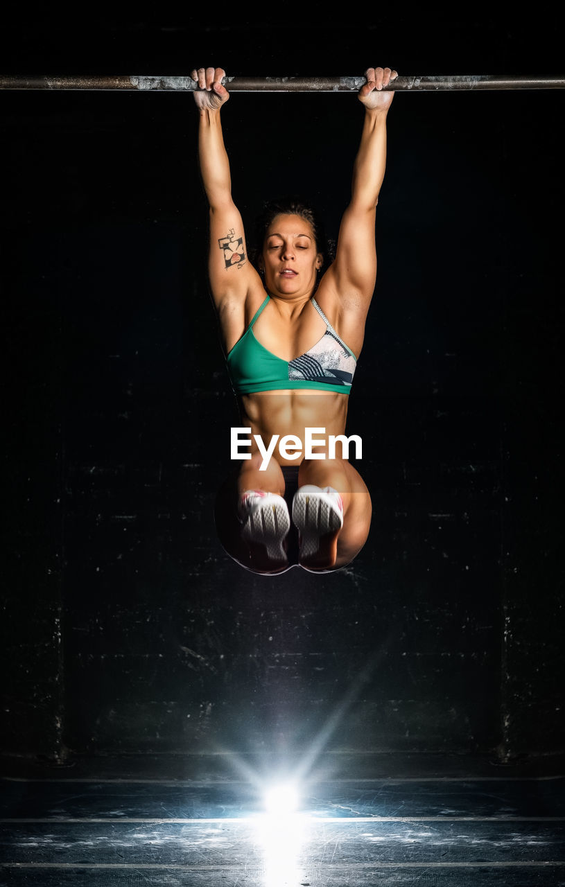 Full body muscular female in sportswear hanging on bar and lifting legs during functional workout in dark gym
