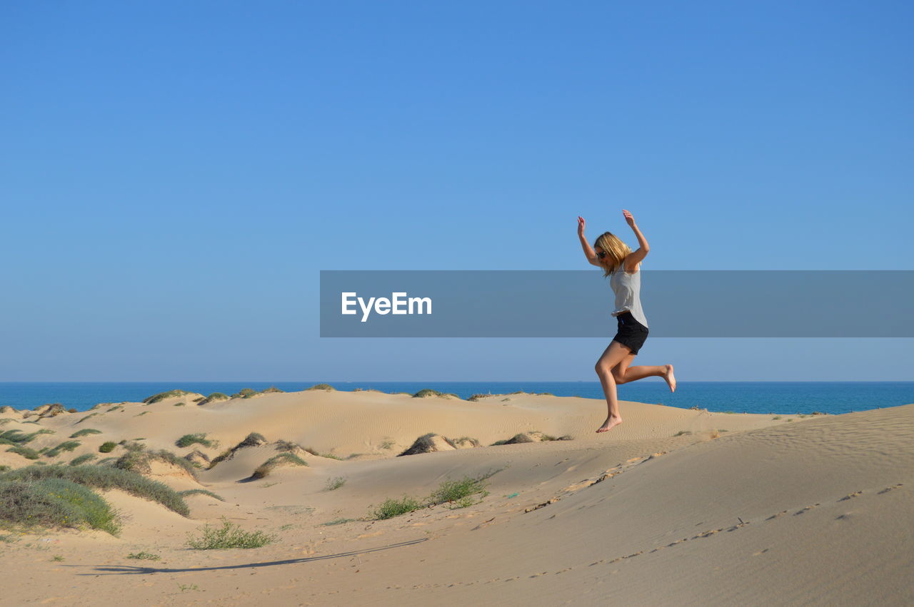 Full length of happy woman jumping on sand at beach against clear blue sky