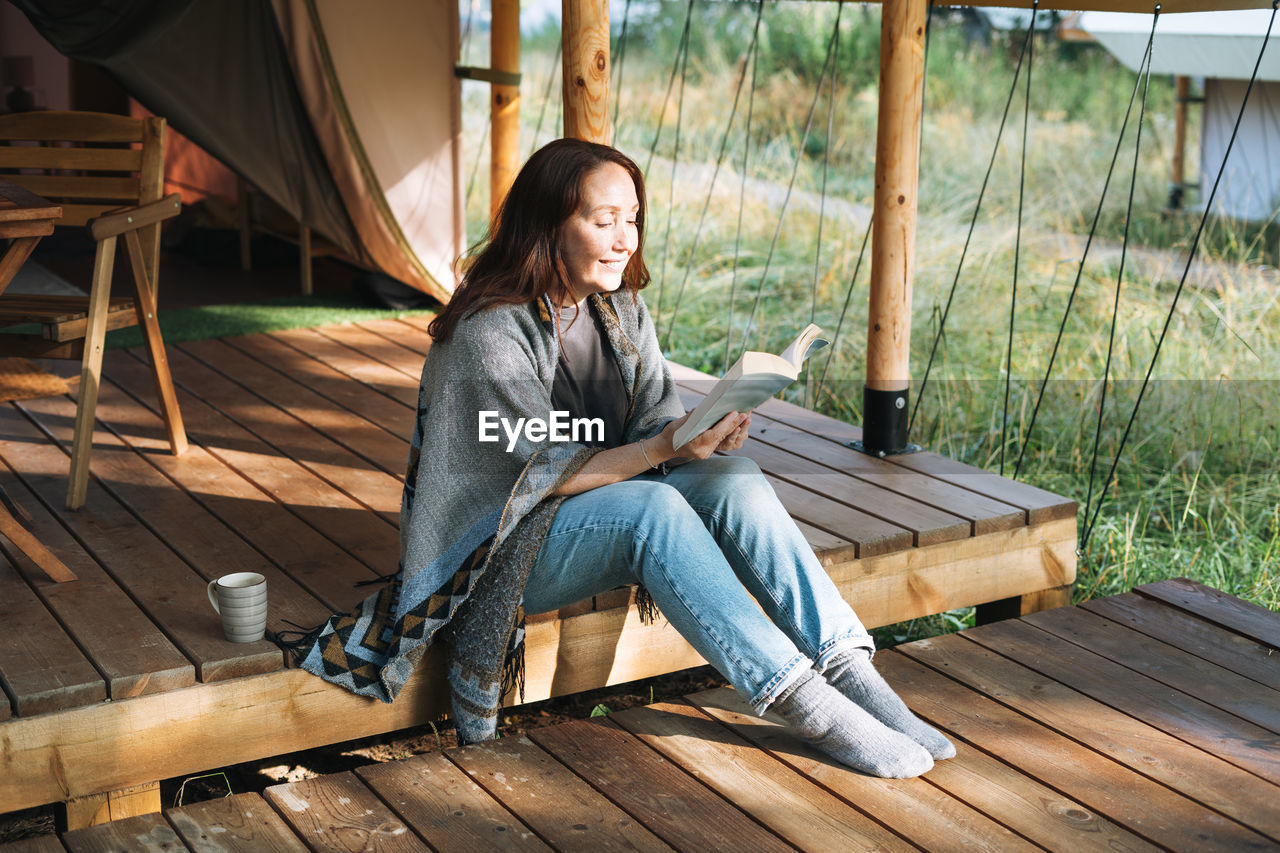 Young brunette woman in poncho reading book and relaxing in glamping in the woods