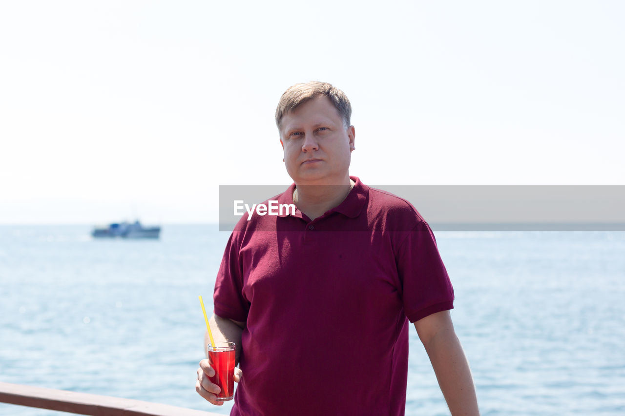 An adult blond man in a bright burgundy t-shirt with a glass of strawberry juice is standing